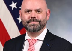 A portrait of ISAC Ericsson, a bald white man with a short, dark beard wearing a dark suit, white shirt, and patterned red tie. He faces forward with a neutral expression in front of an American flag. Integrated Sensing and Communication (ISAC) Advances from Labs to Real-World Trials in 5G/6G Networks